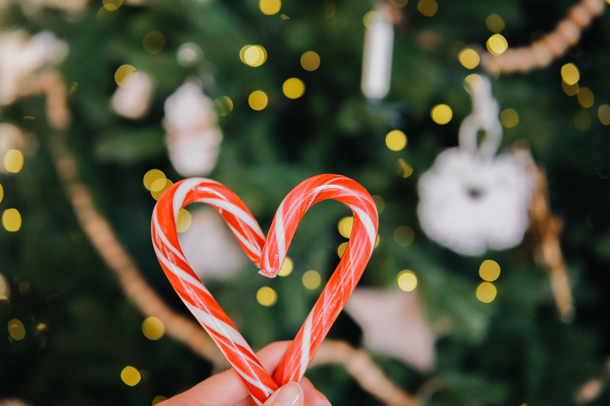 A hand holds a heart-shaped candy cane in front of a Christmas tree adorned with lights and ornaments, capturing the festive spirit. The background is softly blurred, emphasizing the candy.