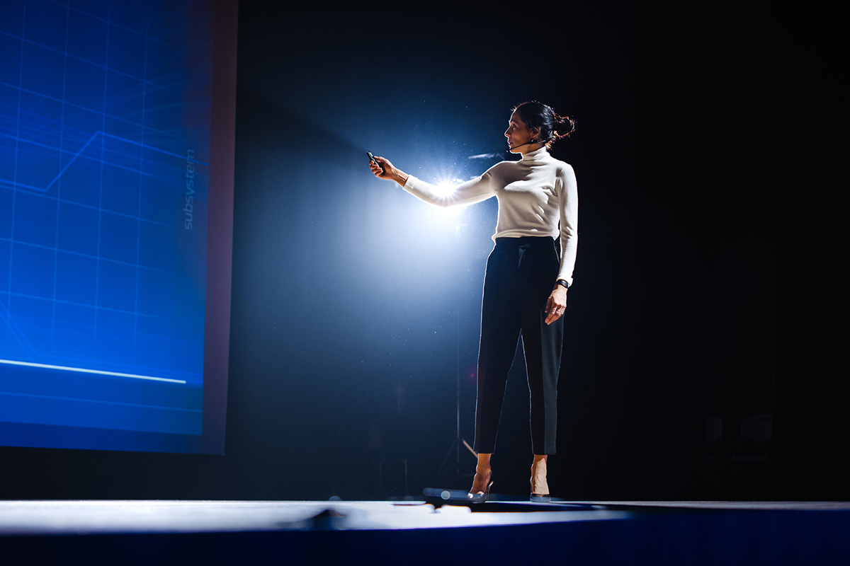 Woman in white shirt and dark pants speaking on a stage in a dark room