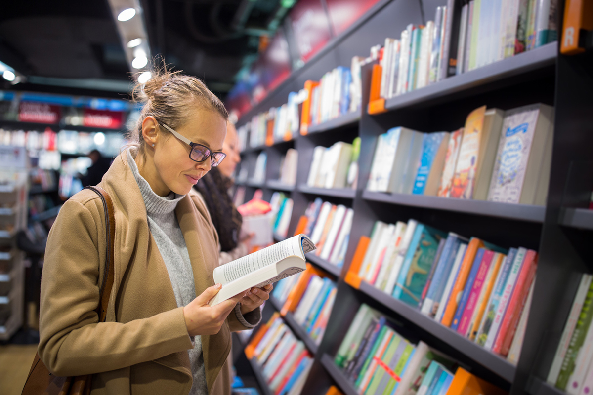 Young white woman in a bookstore reading a book off the shelf.