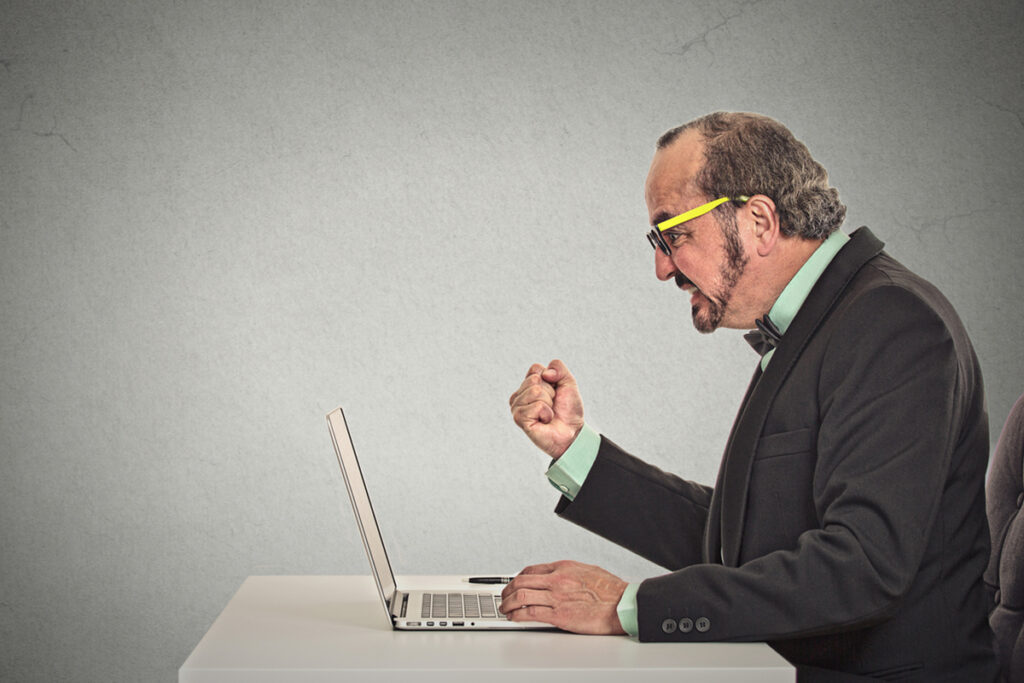 Angry white man wearing glasses, and with a raised fist, yelling at a laptop