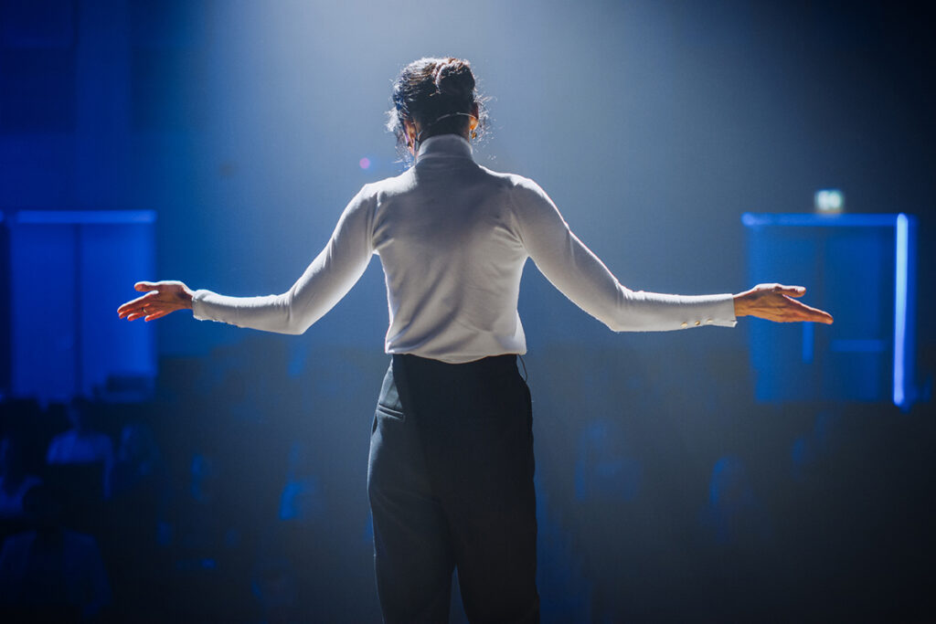 Woman wearing white shirt & dark pants speaking on event stage