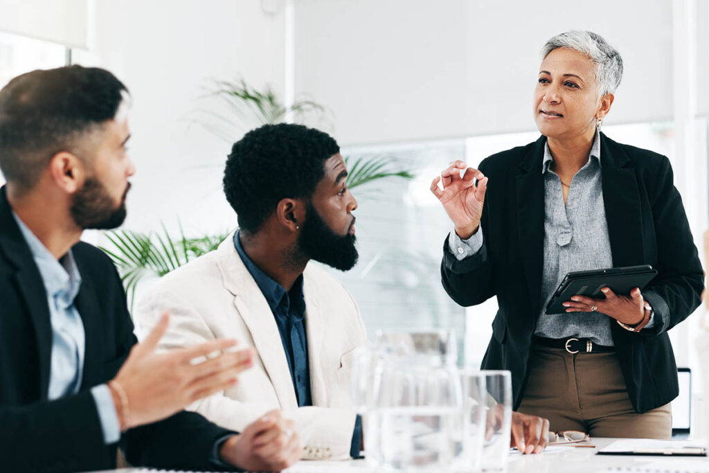 Older female leader in a meeting with her team that includes a young black man and a young white man. The men are paying close attention to what she says. The photo illustrates using storytelling in leadership.