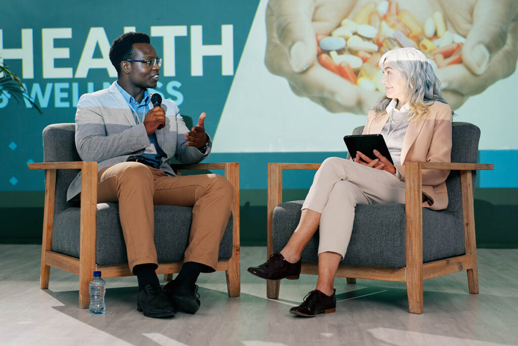 Black man and older white woman sitting in chairs facing each other on stage, illustrating a fireside chat format at a conference.