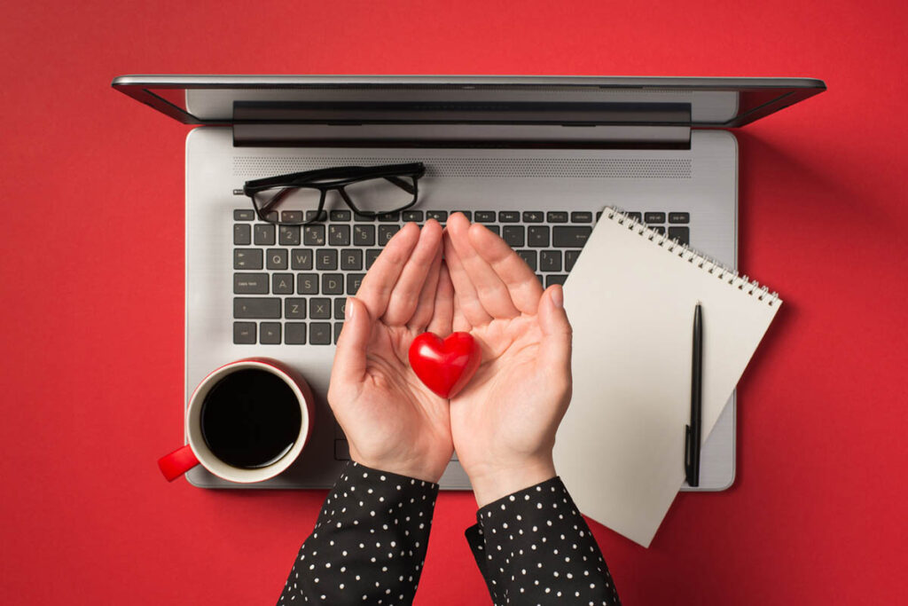 Overhead photo of grey laptop notepad pen glasses cup of coffee and hands holding a red heart isolated on the red backdrop, representing finding meaning in work