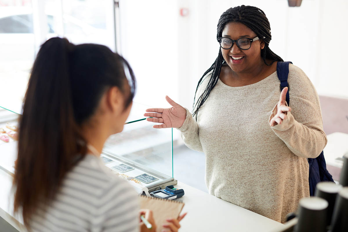 Young female shop owner and young black female customer smiling across the counter, illustrating how special requests can build positive partnerships.