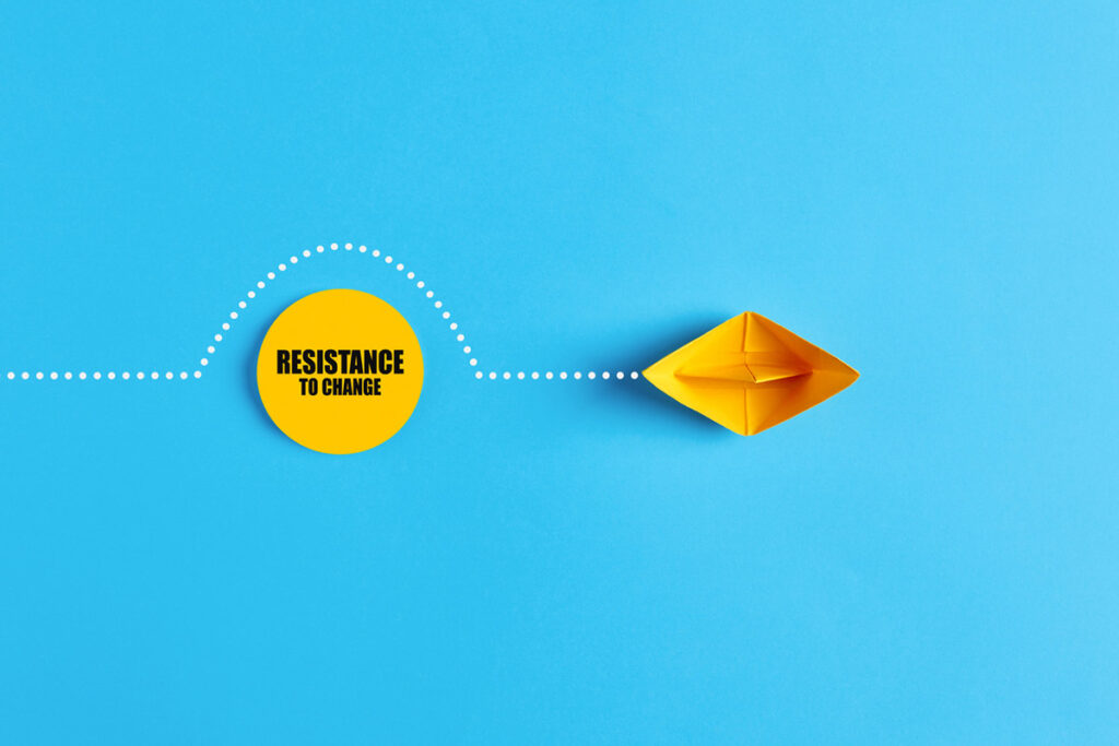 Yellow paper boat navigating around a yellow circle labeled “Resistance to Change” on a blue background, symbolizing overcoming resistance to change.