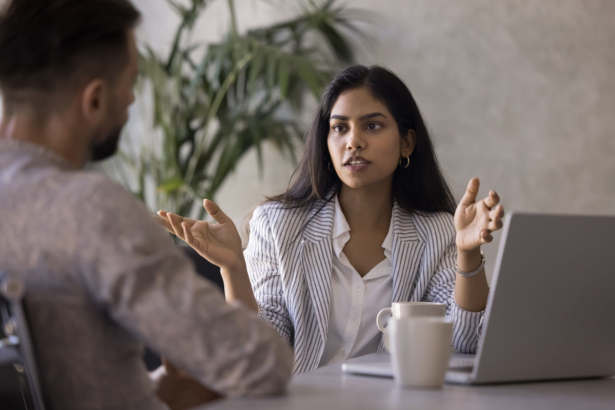A young woman with long dark hair and dark skin sits at a desk with a laptop, discussing a project with a client. The image represents advisor accountability and collaboration in action.