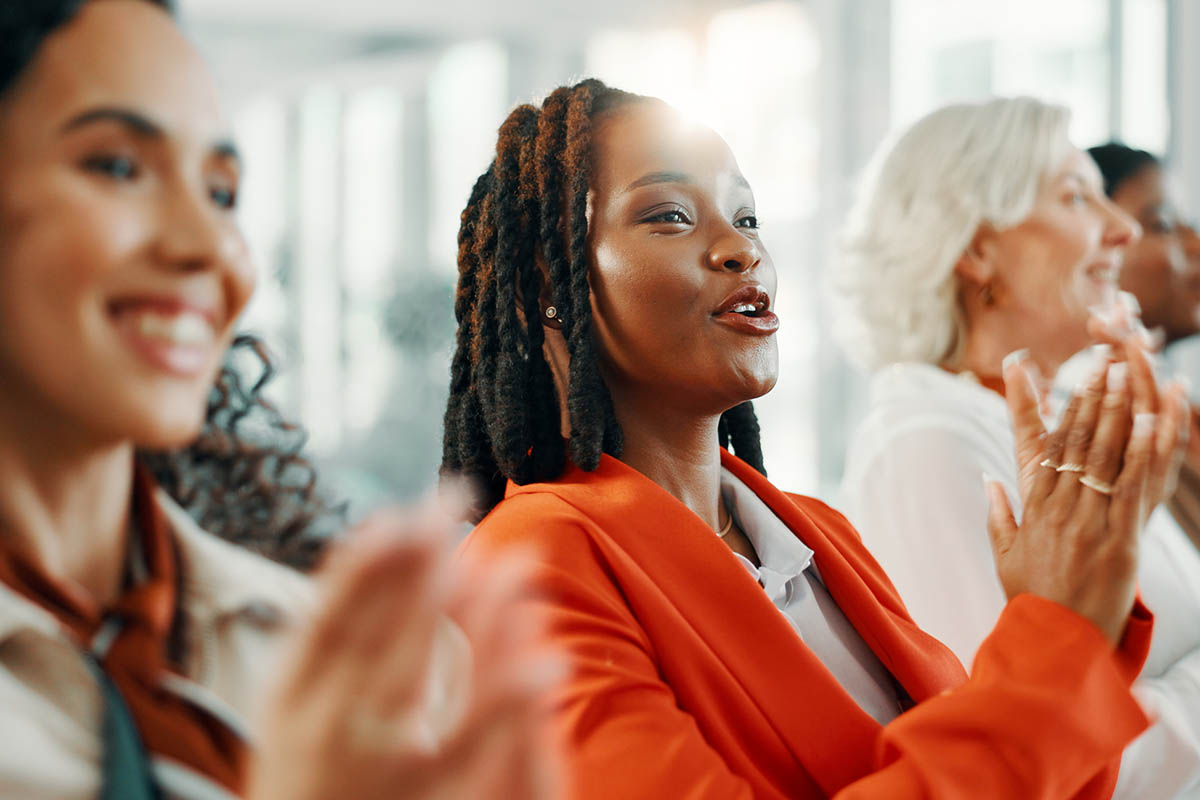 Three women seated in the audience at a professional event applaud during a presentation. The woman in the foreground is Black, wears an orange blazer and has shoulder-length hair, while two women beside her smile and clap in a bright conference space, illustrating audience response during peak experience
