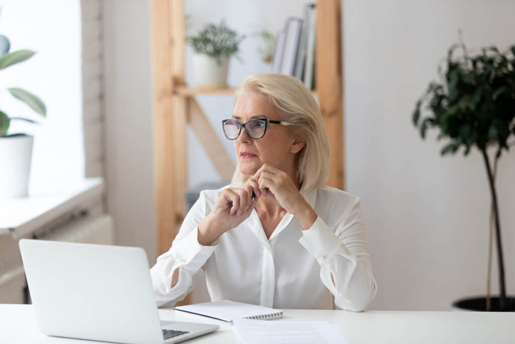 An older professional woman with short gray hair and glasses sits at a desk with a laptop and notebook, resting her hands near her face while looking thoughtfully to the side, illustrating a moment of pause during emotional decision making.