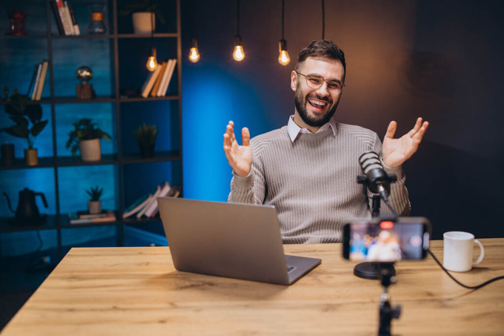 A man with glasses and a beard sits at a desk in a home studio, smiling and gesturing while recording a video with a microphone, laptop, and smartphone on a tripod, illustrating a modern influencer marketing strategy focused on content creation and audience engagement.