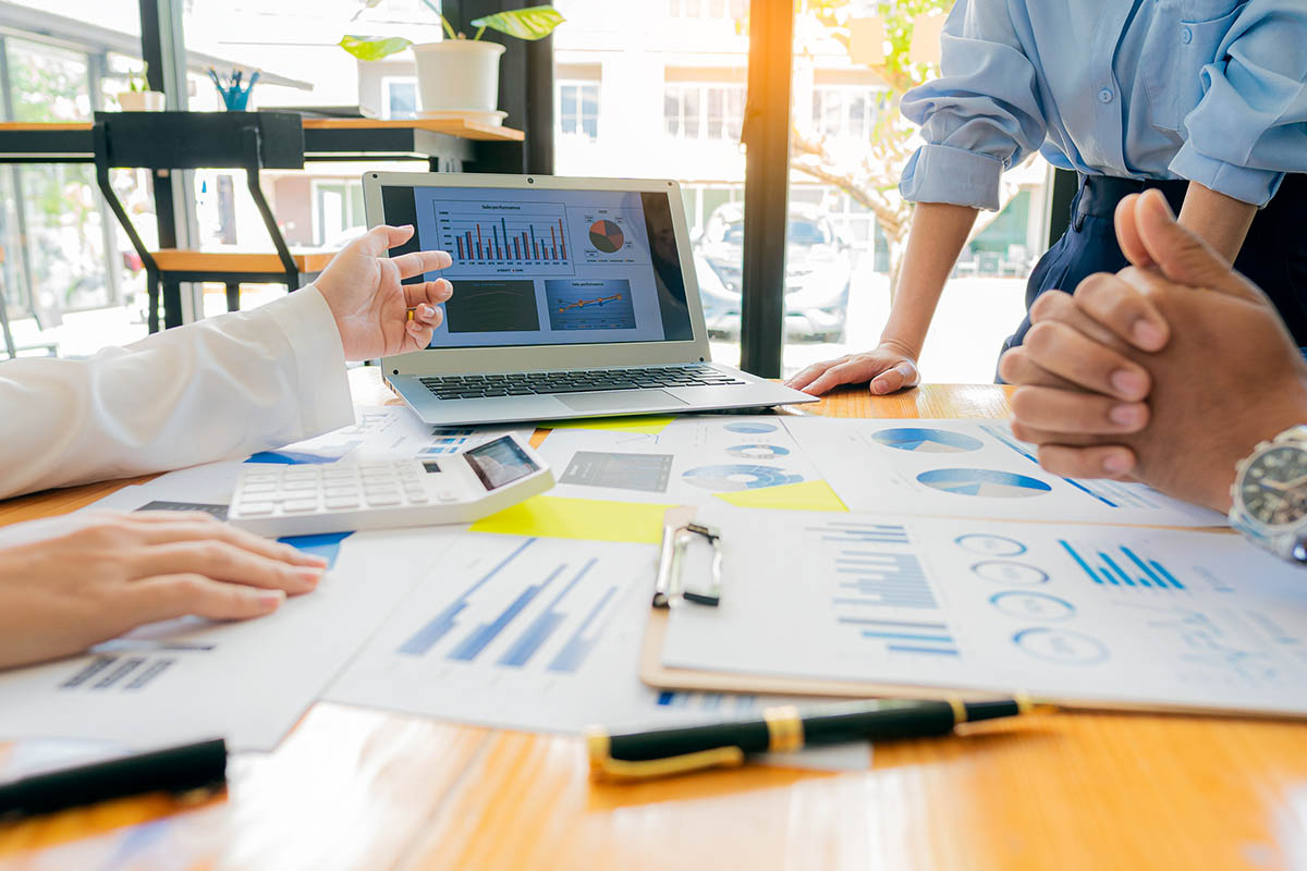 Three business professionals review charts and financial reports on a laptop and printed documents during a meeting, illustrating decision-making and planning for a sales strategy in a volatile market.