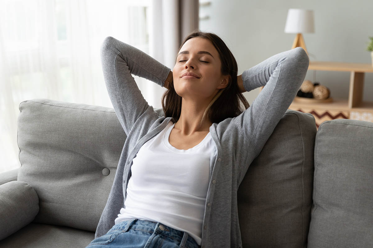 A woman sitting on a gray sofa with her eyes closed and hands behind her head during a quiet afternoon break, illustrating the power of walking away.
