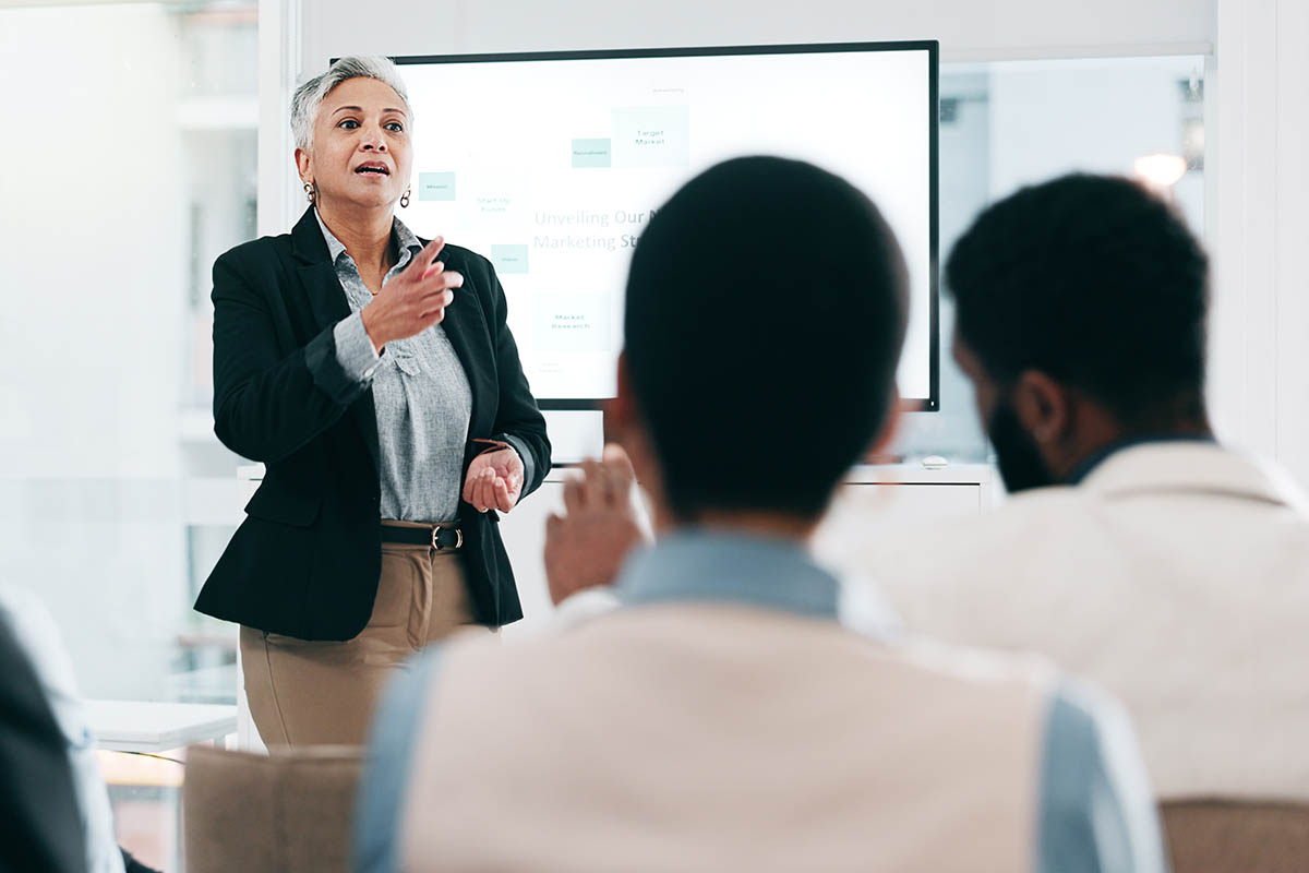 Confident older business leader with short gray hair presenting to a seated group during a meeting, illustrating why buyers must trust the messenger before the message.