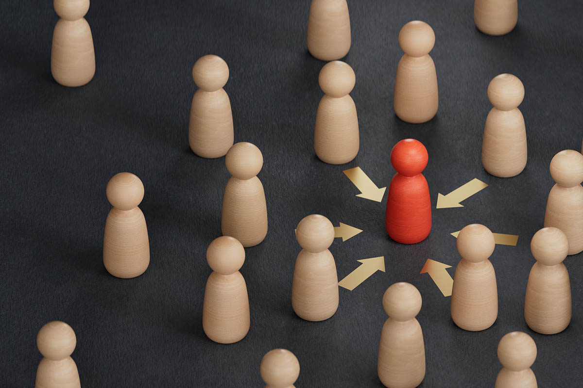 Wooden figures surrounding a single red figure with arrows pointing toward it, symbolizing how to attract ideal clients and stand out in a crowded market.