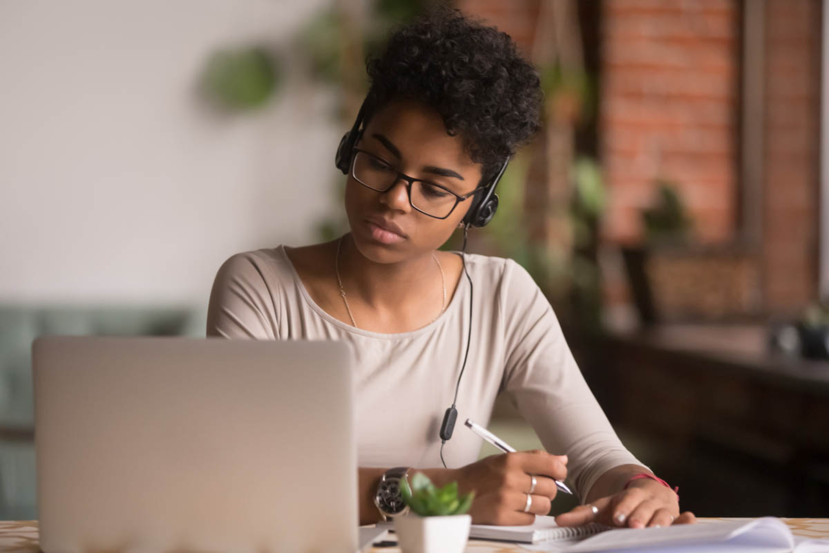 A young black woman with tan skin, wearing a beige shirt and her hair pulled up, researching on a laptop and taking notes, representing a shift in B2B buyer behavior and its impact on outbound B2B marketing.