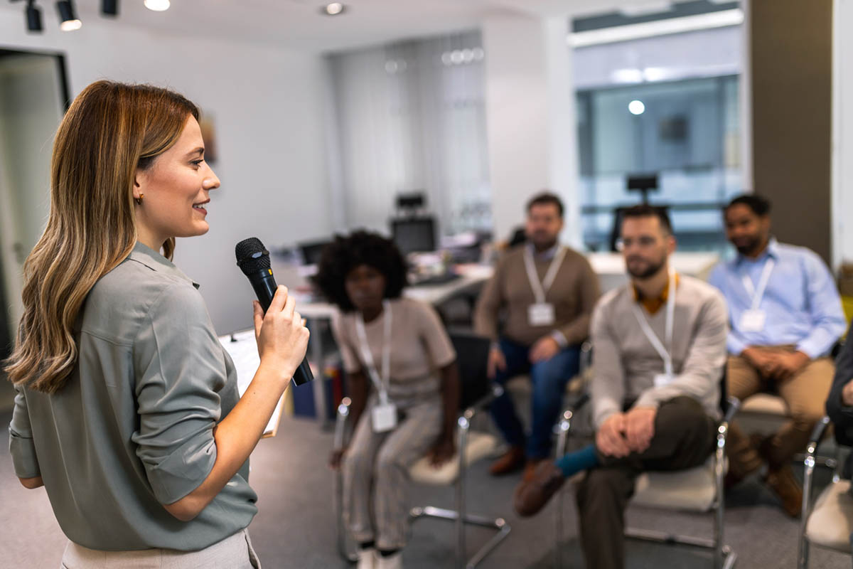 A woman holding a microphone speaks to a small seated group in a professional setting, suggesting expert-led discussion and B2B influencer partnerships.