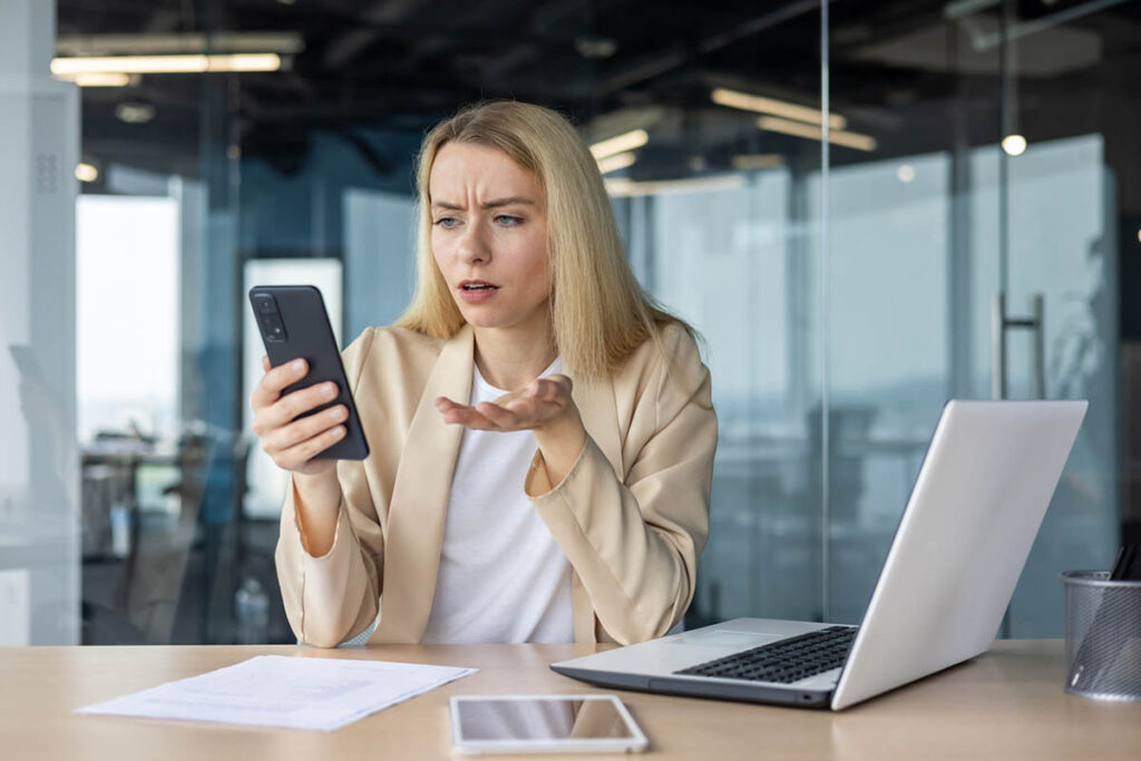 A young white woman with long blonde hair, wearing a white shirt and a beige sweater, in an office looks at her phone with a puzzled expression while sitting at a desk with a laptop and papers, suggesting she is reacting to a message or comment and wondering how to respond to criticism on social media.