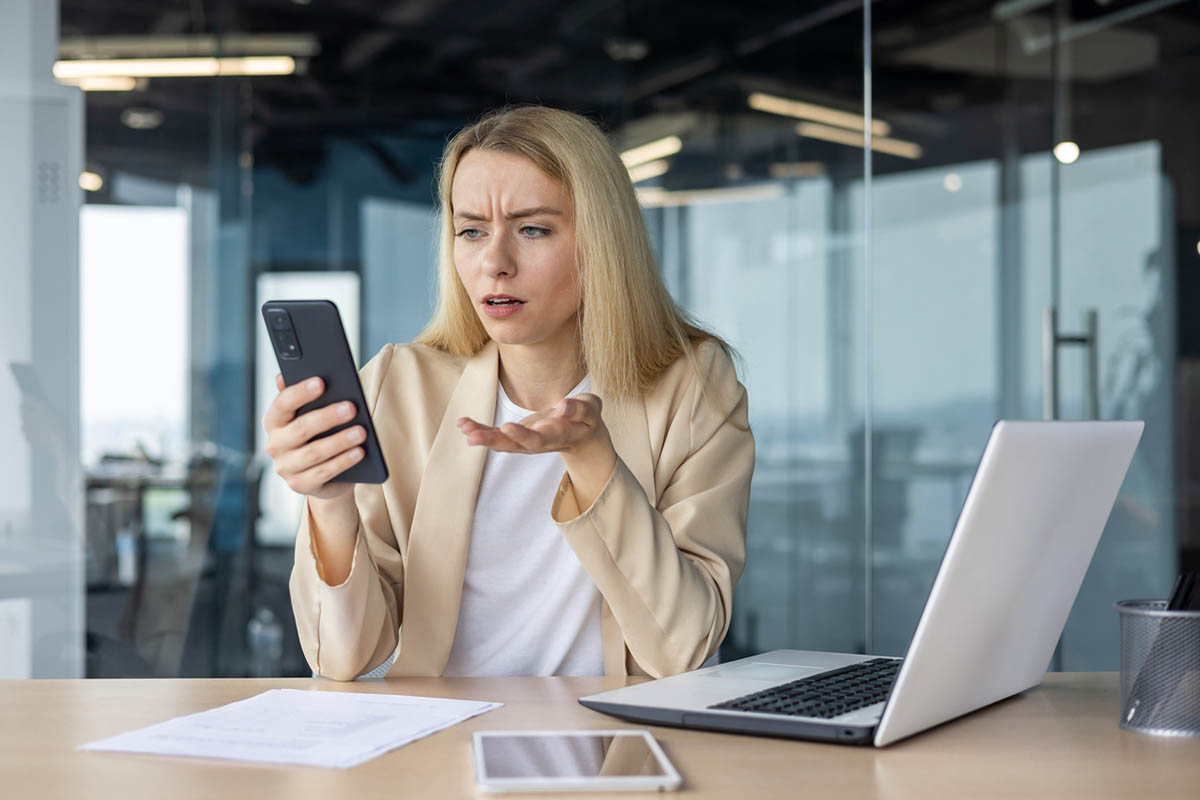 A young white woman with long blonde hair, wearing a white shirt and a beige sweater, in an office looks at her phone with a puzzled expression while sitting at a desk with a laptop and papers, suggesting she is reacting to a message or comment and wondering how to respond to criticism on social media.