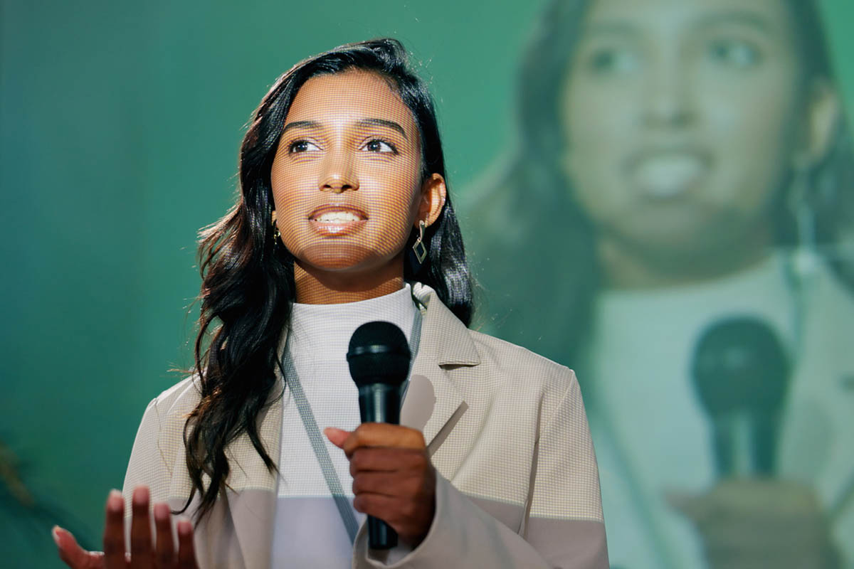 A young professional woman with brown hair pulled back speaks on stage, holding a microphone and wearing a light-colored blazer, demonstrating a strong thought leadership voice and confident presence.