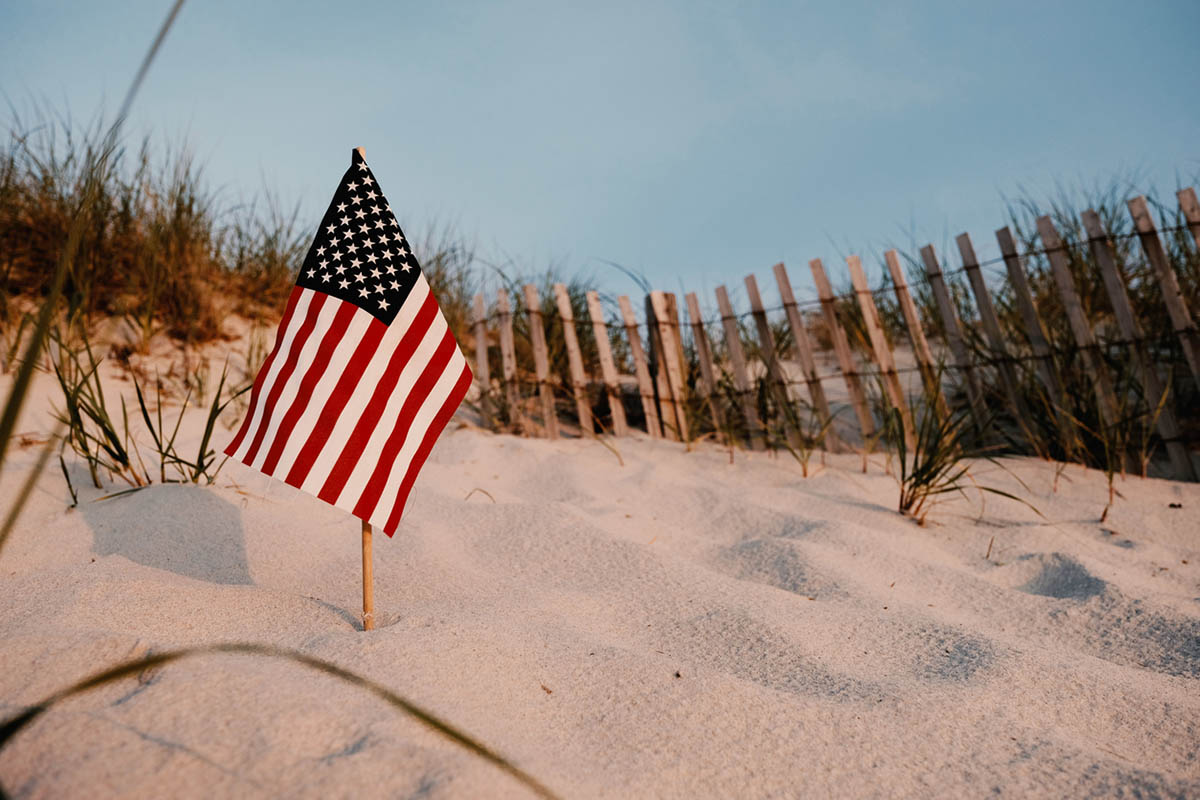 American flag planted in beach sand beside dune grass and a wooden fence at sunset, symbolizing remembrance and gratitude in this Memorial Day reflection.
