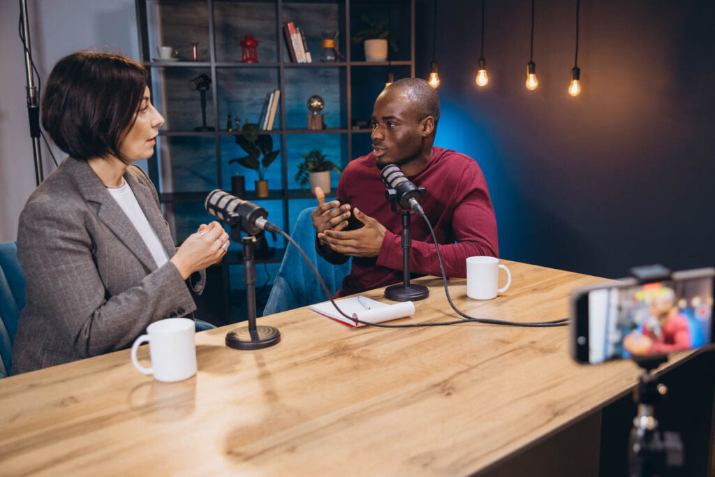 Two podcasters sit across from each other in a modern recording studio, speaking into microphones during a conversational interview, illustrating building audience trust one conversation at a time. A smartphone records the discussion while coffee mugs, notebooks, and studio lighting create an authentic podcast setting.