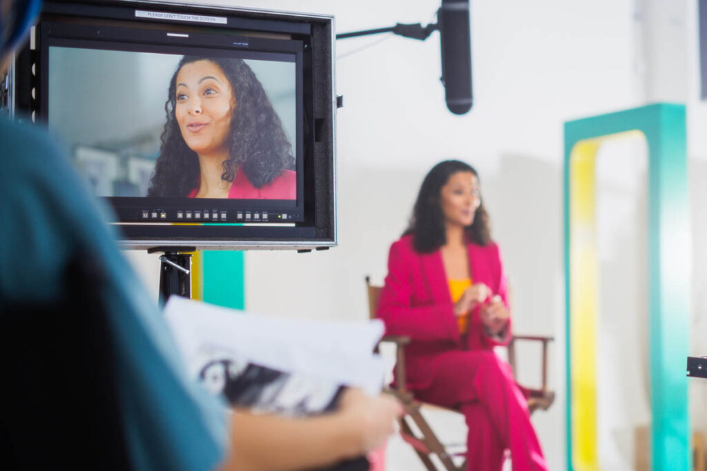 A woman with long dark hair and wearing a red suit records a video in a professional studio, illustrating one of the best tactics to spread marketing messages: customer stories shared with other people.