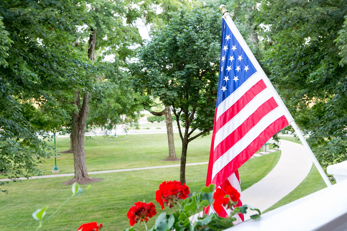 An American flag hangs from a front porch overlooking a quiet, tree-lined neighborhood, symbolizing the moments of freedom we celebrate on Independence Day and the everyday choices they represent.