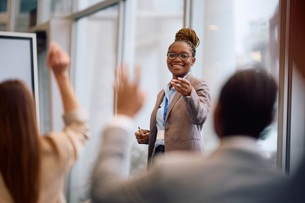 Smiling Black businesswoman wearing glasses and a tan blazer leads an interactive leadership training session as attendees raise their hands, illustrating training soft skills for leaders.