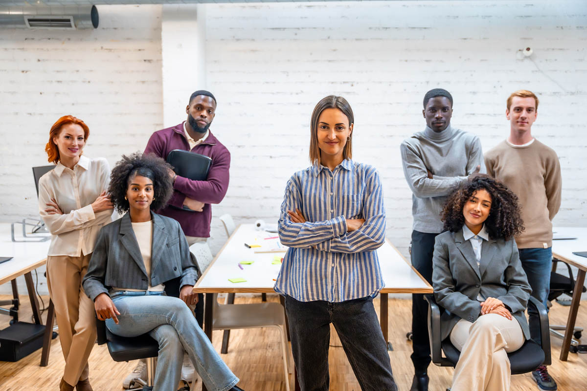A diverse group of seven professionals poses together in a modern office, with a woman standing confidently at the center and colleagues seated and standing around her, representing the importance of communicating with Gen Z leaders in today's collaborative workplace.