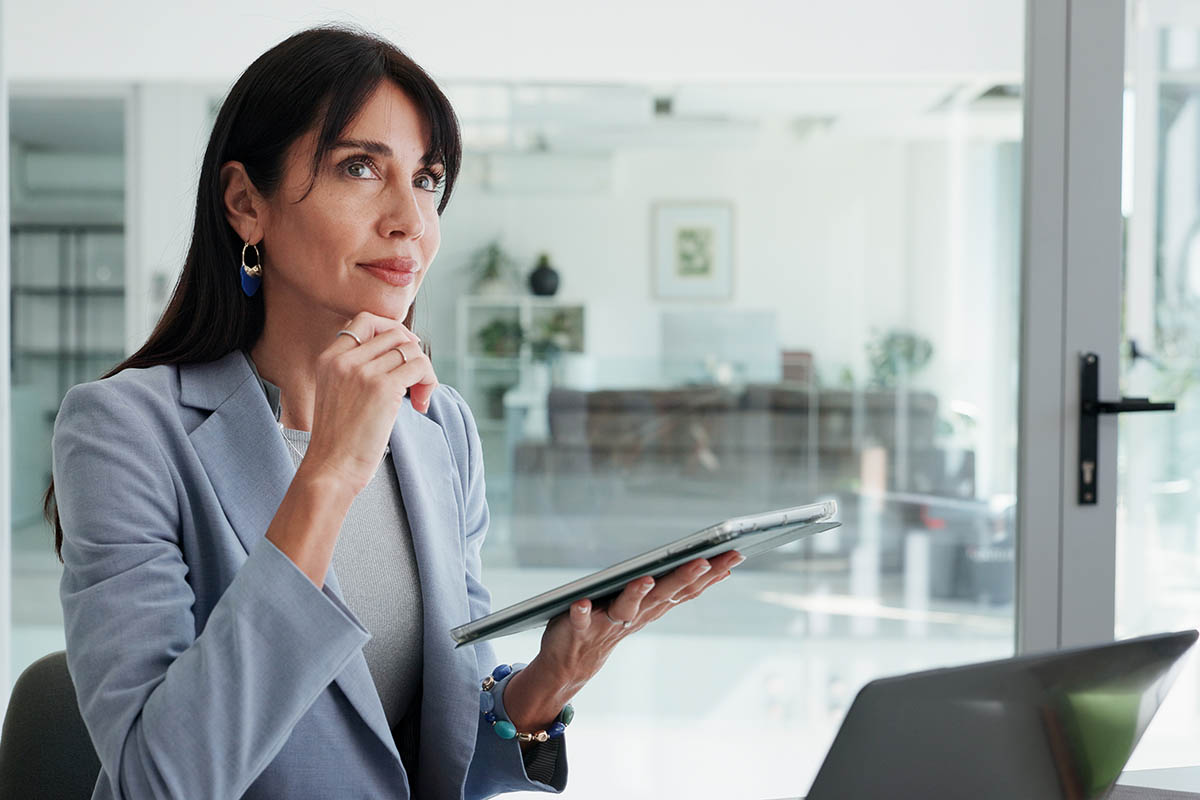 Businesswoman holding a tablet and thoughtfully looking into the distance while working at her desk, illustrating the careful decision-making behind a successful innovation strategy.