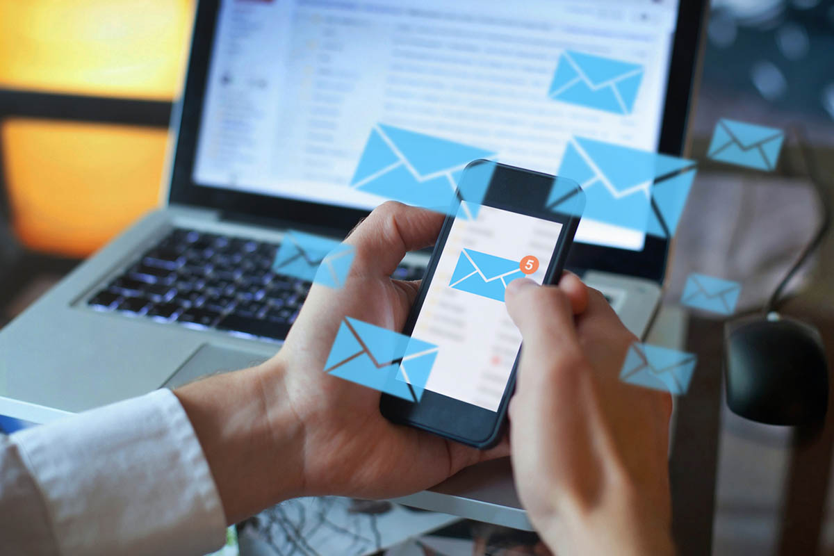 Close-up of a professional checking emails on a smartphone while working on a laptop, with illustrated email icons floating around the devices to represent outbound marketing and digital outreach.