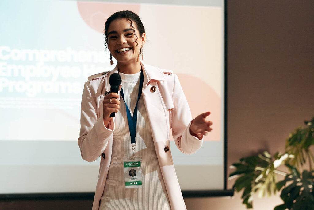 Smiling woman with dark curly hair, wearing a light pink blazer and event badge, holds a microphone while speaking in front of a presentation screen at a professional conference, where experts may get paid to speak.