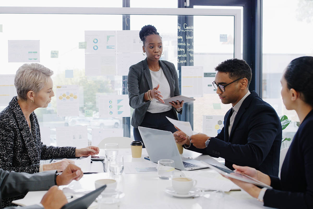A Black woman in a gray blazer stands at a conference table presenting to a diverse group of business executives. An older white woman with short gray hair and a man in a dark suit and glasses listen as she speaks, while other colleagues sit around the table with laptops and documents. The scene illustrates an advisor selling to executives during a business presentation.