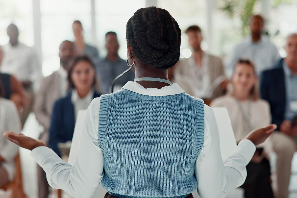 Rear view of a Black woman with braided hair speaking to a diverse audience at a business event, illustrating changing event speaker expectations and audience engagement.