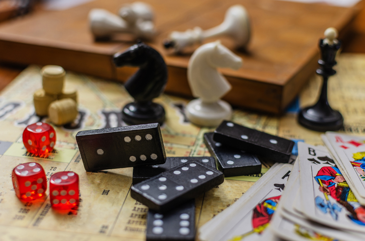 A close-up still life of various classic board games. The scene features a collection of leisure and hobby items including dominoes, red dice, playing cards and chess pieces.